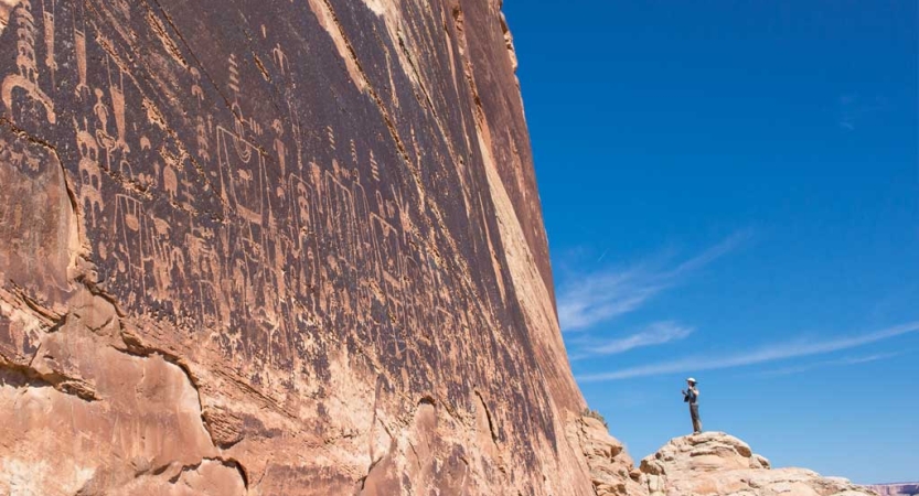 A person stands beside a tall canyon wall, looking up at ancient markings. 